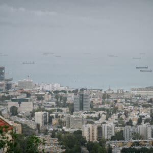 חיפה, נמל חיפה, מפרץ חיפה, הים התיכון, אוניות משא, ספינות, קו חוף, עיר חוף, נוף עירוני, מבנים, בניינים גבוהים, שכונות מגורים, אזור תעשייה, שמיים מעוננים, מזג אוויר חורפי, ערפל ימי, אובך, שמיים אפורים, זיהום אוויר, ראות נמוכה
