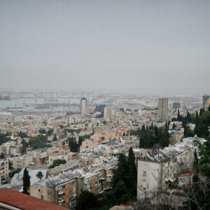 חיפה, נמל חיפה, מפרץ חיפה, הים התיכון, אוניות משא, ספינות, קו חוף, עיר חוף, נוף עירוני, מבנים, בניינים גבוהים, שכונות מגורים, אזור תעשייה, שמיים מעוננים, מזג אוויר חורפי, ערפל ימי, אובך, שמיים אפורים, זיהום אוויר, ראות נמוכה