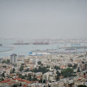 חיפה, נמל חיפה, מפרץ חיפה, הים התיכון, אוניות משא, ספינות, קו חוף, עיר חוף, נוף עירוני, מבנים, בניינים גבוהים, שכונות מגורים, אזור תעשייה, שמיים מעוננים, מזג אוויר חורפי, ערפל ימי, אובך, שמיים אפורים, זיהום אוויר, ראות נמוכה