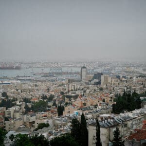 חיפה, נמל חיפה, מפרץ חיפה, הים התיכון, אוניות משא, ספינות, קו חוף, עיר חוף, נוף עירוני, מבנים, בניינים גבוהים, שכונות מגורים, אזור תעשייה, שמיים מעוננים, מזג אוויר חורפי, ערפל ימי, אובך, שמיים אפורים, זיהום אוויר, ראות נמוכה