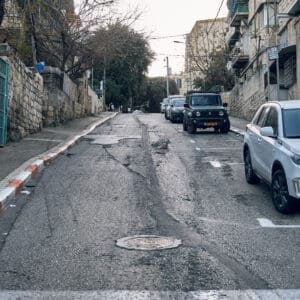 רחוב סירקין, שוק תלפיות, שכונת הדר, רחוב עירוני, כביש סדוק, אספלט בלוי, מכסה ביוב, מדרכה צרה, אבני שפה אדום לבן, חניה ברחוב, רכבים חונים, קיר אבן ישן, גדר מתכת, בנייני מגורים ותיקים, סורגים לחלונות, מרפסות קטנות, תשתיות חשופות, עץ עירוני, עלייה תלולה, תיעוד עירוני
