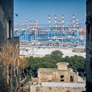 ואדי סליב, סאליב, נמל חיפה, עגורני נמל, מנופי גשר, מסוף מכולות, מכולות מטען, מפרץ חיפה, תעשייה ימית, קו רקיע תעשייתי, בתי אבן ישנים, אדריכלות עותמאנית, מרפסות ברזל, חלונות מקושתים, טופוגרפיה הררית, מבט דרך שכונה היסטורית, ניגוד ישן וחדש, מרקם עירוני, מורשת אדריכלית, לוגיסטיקה ימית, צילום נוף עירוני