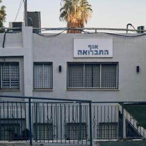 אגף התברואה חיפה, עיריית חיפה, מבנה משרדי עירוני, שירותי תברואה, פינוי אשפה, תחזוקה עירונית, בניין תפעולי, חזית בטון אפורה, סורגים לחלונות, שילוט עברי, מתחם עירוני, חצר פנימית, תשתיות חשמל חשופות, משרדי מנהלה, שירות לתושב, מבנה ציבורי, צילום תיעודי, תפעול עירוני
