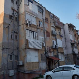 פינוי בינוי, התחדשות עירונית, תמ״א 38, הריסה ובנייה מחדש, חיזוק מבנים, שדרוג תשתיות, בנייה רוויה, צפיפות עירונית, תכנון עירוני, מדיניות דיור, התחדשות שכונות ותיקות, פיתוח עירוני, שינוי ייעוד קרקע, הגדלת זכויות בנייה, שימור מול פיתוח, חידוש חזיתות, שיפור איכות חיים, השקעות נדל״ן, עליית ערך נכסים, פרויקטים למגורים, שיתוף דיירים, פיצוי דיירים, יזמות נדל״ן, רגולציה תכנונית, היתרי בנייה, ועדות תכנון ובנייה, שיקום מבנים ישנים, התחדשות תשתיות עירוניות, התחדשות מרכזי ערים, בנייה לגובה, בניין מגורים ישן, שיכון, בלוק דירות, חזית בניין, ארכיטקטורה ישראלית, שכונת מגורים, עיר ישראלית, רחוב עירוני, תחנת אוטובוס, תחבורה ציבורית, תחנת המתנה, עץ אורן, צמחייה עירונית, מדרכה, אבני סלע, כביש רטוב, שמש חזקה, שמיים כחולים, אור וצל, חזית מוזנחת, תריסים סגורים, מזגנים חיצוניים, אנטנות על הגג, צילום רחוב, סביבה עירונית, דיור ציבורי, מבנה בטון, שכונה ותיקה, נוף עירוני