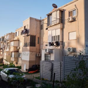 פינוי בינוי, התחדשות עירונית, תמ״א 38, הריסה ובנייה מחדש, חיזוק מבנים, שדרוג תשתיות, בנייה רוויה, צפיפות עירונית, תכנון עירוני, מדיניות דיור, התחדשות שכונות ותיקות, פיתוח עירוני, שינוי ייעוד קרקע, הגדלת זכויות בנייה, שימור מול פיתוח, חידוש חזיתות, שיפור איכות חיים, השקעות נדל״ן, עליית ערך נכסים, פרויקטים למגורים, שיתוף דיירים, פיצוי דיירים, יזמות נדל״ן, רגולציה תכנונית, היתרי בנייה, ועדות תכנון ובנייה, שיקום מבנים ישנים, התחדשות תשתיות עירוניות, התחדשות מרכזי ערים, בנייה לגובה, בניין מגורים ישן, שיכון, בלוק דירות, חזית בניין, ארכיטקטורה ישראלית, שכונת מגורים, עיר ישראלית, רחוב עירוני, תחנת אוטובוס, תחבורה ציבורית, תחנת המתנה, עץ אורן, צמחייה עירונית, מדרכה, אבני סלע, כביש רטוב, שמש חזקה, שמיים כחולים, אור וצל, חזית מוזנחת, תריסים סגורים, מזגנים חיצוניים, אנטנות על הגג, צילום רחוב, סביבה עירונית, דיור ציבורי, מבנה בטון, שכונה ותיקה, נוף עירוני