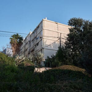 פינוי בינוי, התחדשות עירונית, תמ״א 38, הריסה ובנייה מחדש, חיזוק מבנים, שדרוג תשתיות, בנייה רוויה, צפיפות עירונית, תכנון עירוני, מדיניות דיור, התחדשות שכונות ותיקות, פיתוח עירוני, שינוי ייעוד קרקע, הגדלת זכויות בנייה, שימור מול פיתוח, חידוש חזיתות, שיפור איכות חיים, השקעות נדל״ן, עליית ערך נכסים, פרויקטים למגורים, שיתוף דיירים, פיצוי דיירים, יזמות נדל״ן, רגולציה תכנונית, היתרי בנייה, ועדות תכנון ובנייה, שיקום מבנים ישנים, התחדשות תשתיות עירוניות, התחדשות מרכזי ערים, בנייה לגובה, בניין מגורים ישן, שיכון, בלוק דירות, חזית בניין, ארכיטקטורה ישראלית, שכונת מגורים, עיר ישראלית, רחוב עירוני, תחנת אוטובוס, תחבורה ציבורית, תחנת המתנה, עץ אורן, צמחייה עירונית, מדרכה, אבני סלע, כביש רטוב, שמש חזקה, שמיים כחולים, אור וצל, חזית מוזנחת, תריסים סגורים, מזגנים חיצוניים, אנטנות על הגג, צילום רחוב, סביבה עירונית, דיור ציבורי, מבנה בטון, שכונה ותיקה, נוף עירוני