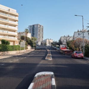 כביש ריק, רחוב שומם, עיר ריקה, היעדר תנועה, חירום לאומי, מצב חירום, מבצע שאגת הארי, מתיחות ביטחונית, עורף אזרחי, שגרה מופרעת, תחנת אוטובוס ריקה, תחבורה ציבורית מושבתת, בנייני מגורים ישנים, מגדל מגורים חדש, התחדשות עירונית, פינוי בינוי, שכונה ותיקה, שמיים בהירים, יום שמשי, תשתיות עירוניות, כביש עירוני, סימוני דרך, מדרכה ריקה, דממה עירונית, שקט מתוח, חזיתות מבנים, צילום תיעודי, צילום עיתונאי, מציאות ביטחונית בישראל