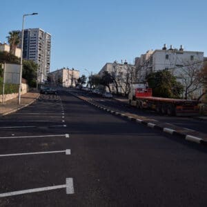 כביש ריק, רחוב שומם, עיר ריקה, היעדר תנועה, חירום לאומי, מצב חירום, מבצע שאגת הארי, מתיחות ביטחונית, עורף אזרחי, שגרה מופרעת, תחנת אוטובוס ריקה, תחבורה ציבורית מושבתת, בנייני מגורים ישנים, מגדל מגורים חדש, התחדשות עירונית, פינוי בינוי, שכונה ותיקה, שמיים בהירים, יום שמשי, תשתיות עירוניות, כביש עירוני, סימוני דרך, מדרכה ריקה, דממה עירונית, שקט מתוח, חזיתות מבנים, צילום תיעודי, צילום עיתונאי, מציאות ביטחונית בישראל