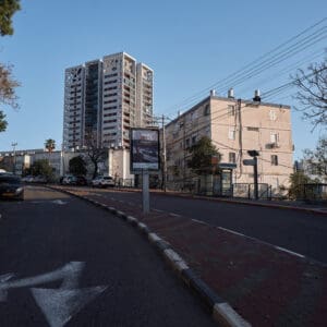 כביש ריק, רחוב שומם, עיר ריקה, היעדר תנועה, חירום לאומי, מצב חירום, מבצע שאגת הארי, מתיחות ביטחונית, עורף אזרחי, שגרה מופרעת, תחנת אוטובוס ריקה, תחבורה ציבורית מושבתת, בנייני מגורים ישנים, מגדל מגורים חדש, התחדשות עירונית, פינוי בינוי, שכונה ותיקה, שמיים בהירים, יום שמשי, תשתיות עירוניות, כביש עירוני, סימוני דרך, מדרכה ריקה, דממה עירונית, שקט מתוח, חזיתות מבנים, צילום תיעודי, צילום עיתונאי, מציאות ביטחונית בישראל, תמ״א 38, הריסה ובנייה מחדש, חיזוק מבנים, שדרוג תשתיות, בנייה רוויה, צפיפות עירונית, תכנון עירוני, מדיניות דיור, התחדשות שכונות ותיקות, פיתוח עירוני, שינוי ייעוד קרקע, הגדלת זכויות בנייה, שימור מול פיתוח, חידוש חזיתות, שיפור איכות חיים, השקעות נדל״ן, עליית ערך נכסים, פרויקטים למגורים, שיתוף דיירים, פיצוי דיירים, יזמות נדל״ן, רגולציה תכנונית, היתרי בנייה, ועדות תכנון ובנייה, שיקום מבנים ישנים, התחדשות תשתיות עירוניות, התחדשות מרכזי ערים, בנייה לגובה, בניין מגורים ישן, שיכון, בלוק דירות, חזית בניין, ארכיטקטורה ישראלית, שכונת מגורים, עיר ישראלית, רחוב עירוני, תחנת אוטובוס, תחבורה ציבורית, תחנת המתנה, עץ אורן, צמחייה עירונית, מדרכה, אבני סלע, כביש רטוב, שמש חזקה, שמיים כחולים, אור וצל, חזית מוזנחת, תריסים סגורים, מזגנים חיצוניים, אנטנות על הגג, צילום רחוב, סביבה עירונית, דיור ציבורי, מבנה בטון, נוף עירוני