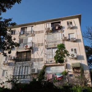 פינוי בינוי, התחדשות עירונית, תמ״א 38, הריסה ובנייה מחדש, חיזוק מבנים, שדרוג תשתיות, בנייה רוויה, צפיפות עירונית, תכנון עירוני, מדיניות דיור, התחדשות שכונות ותיקות, פיתוח עירוני, שינוי ייעוד קרקע, הגדלת זכויות בנייה, שימור מול פיתוח, חידוש חזיתות, שיפור איכות חיים, השקעות נדל״ן, עליית ערך נכסים, פרויקטים למגורים, שיתוף דיירים, פיצוי דיירים, יזמות נדל״ן, רגולציה תכנונית, היתרי בנייה, ועדות תכנון ובנייה, שיקום מבנים ישנים, התחדשות תשתיות עירוניות, התחדשות מרכזי ערים, בנייה לגובה, בניין מגורים ישן, שיכון, בלוק דירות, חזית בניין, ארכיטקטורה ישראלית, שכונת מגורים, עיר ישראלית, רחוב עירוני, תחנת אוטובוס, תחבורה ציבורית, תחנת המתנה, עץ אורן, צמחייה עירונית, מדרכה, אבני סלע, כביש רטוב, שמש חזקה, שמיים כחולים, אור וצל, חזית מוזנחת, תריסים סגורים, מזגנים חיצוניים, אנטנות על הגג, צילום רחוב, סביבה עירונית, דיור ציבורי, מבנה בטון, שכונה ותיקה, נוף עירוני