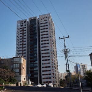 פינוי בינוי, התחדשות עירונית, תמ״א 38, הריסה ובנייה מחדש, חיזוק מבנים, שדרוג תשתיות, בנייה רוויה, צפיפות עירונית, תכנון עירוני, מדיניות דיור, התחדשות שכונות ותיקות, פיתוח עירוני, שינוי ייעוד קרקע, הגדלת זכויות בנייה, שימור מול פיתוח, חידוש חזיתות, שיפור איכות חיים, השקעות נדל״ן, עליית ערך נכסים, פרויקטים למגורים, שיתוף דיירים, פיצוי דיירים, יזמות נדל״ן, רגולציה תכנונית, היתרי בנייה, ועדות תכנון ובנייה, שיקום מבנים ישנים, התחדשות תשתיות עירוניות, התחדשות מרכזי ערים, בנייה לגובה, בניין מגורים ישן, שיכון, בלוק דירות, חזית בניין, ארכיטקטורה ישראלית, שכונת מגורים, עיר ישראלית, רחוב עירוני, תחנת אוטובוס, תחבורה ציבורית, תחנת המתנה, עץ אורן, צמחייה עירונית, מדרכה, אבני סלע, כביש רטוב, שמש חזקה, שמיים כחולים, אור וצל, חזית מוזנחת, תריסים סגורים, מזגנים חיצוניים, אנטנות על הגג, צילום רחוב, סביבה עירונית, דיור ציבורי, מבנה בטון, שכונה ותיקה, נוף עירוני