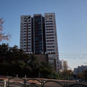 פינוי בינוי, התחדשות עירונית, תמ״א 38, הריסה ובנייה מחדש, חיזוק מבנים, שדרוג תשתיות, בנייה רוויה, צפיפות עירונית, תכנון עירוני, מדיניות דיור, התחדשות שכונות ותיקות, פיתוח עירוני, שינוי ייעוד קרקע, הגדלת זכויות בנייה, שימור מול פיתוח, חידוש חזיתות, שיפור איכות חיים, השקעות נדל״ן, עליית ערך נכסים, פרויקטים למגורים, שיתוף דיירים, פיצוי דיירים, יזמות נדל״ן, רגולציה תכנונית, היתרי בנייה, ועדות תכנון ובנייה, שיקום מבנים ישנים, התחדשות תשתיות עירוניות, התחדשות מרכזי ערים, בנייה לגובה, בניין מגורים ישן, שיכון, בלוק דירות, חזית בניין, ארכיטקטורה ישראלית, שכונת מגורים, עיר ישראלית, רחוב עירוני, תחנת אוטובוס, תחבורה ציבורית, תחנת המתנה, עץ אורן, צמחייה עירונית, מדרכה, אבני סלע, כביש רטוב, שמש חזקה, שמיים כחולים, אור וצל, חזית מוזנחת, תריסים סגורים, מזגנים חיצוניים, אנטנות על הגג, צילום רחוב, סביבה עירונית, דיור ציבורי, מבנה בטון, שכונה ותיקה, נוף עירוני