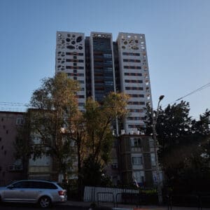 פינוי בינוי, התחדשות עירונית, תמ״א 38, הריסה ובנייה מחדש, חיזוק מבנים, שדרוג תשתיות, בנייה רוויה, צפיפות עירונית, תכנון עירוני, מדיניות דיור, התחדשות שכונות ותיקות, פיתוח עירוני, שינוי ייעוד קרקע, הגדלת זכויות בנייה, שימור מול פיתוח, חידוש חזיתות, שיפור איכות חיים, השקעות נדל״ן, עליית ערך נכסים, פרויקטים למגורים, שיתוף דיירים, פיצוי דיירים, יזמות נדל״ן, רגולציה תכנונית, היתרי בנייה, ועדות תכנון ובנייה, שיקום מבנים ישנים, התחדשות תשתיות עירוניות, התחדשות מרכזי ערים, בנייה לגובה, בניין מגורים ישן, שיכון, בלוק דירות, חזית בניין, ארכיטקטורה ישראלית, שכונת מגורים, עיר ישראלית, רחוב עירוני, תחנת אוטובוס, תחבורה ציבורית, תחנת המתנה, עץ אורן, צמחייה עירונית, מדרכה, אבני סלע, כביש רטוב, שמש חזקה, שמיים כחולים, אור וצל, חזית מוזנחת, תריסים סגורים, מזגנים חיצוניים, אנטנות על הגג, צילום רחוב, סביבה עירונית, דיור ציבורי, מבנה בטון, שכונה ותיקה, נוף עירוני