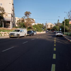 כביש ריק, רחוב שומם, עיר ריקה, היעדר תנועה, חירום לאומי, מצב חירום, מבצע שאגת הארי, מתיחות ביטחונית, עורף אזרחי, שגרה מופרעת, תחנת אוטובוס ריקה, תחבורה ציבורית מושבתת, בנייני מגורים ישנים, מגדל מגורים חדש, התחדשות עירונית, פינוי בינוי, שכונה ותיקה, שמיים בהירים, יום שמשי, תשתיות עירוניות, כביש עירוני, סימוני דרך, מדרכה ריקה, דממה עירונית, שקט מתוח, חזיתות מבנים, צילום תיעודי, צילום עיתונאי, מציאות ביטחונית בישראל