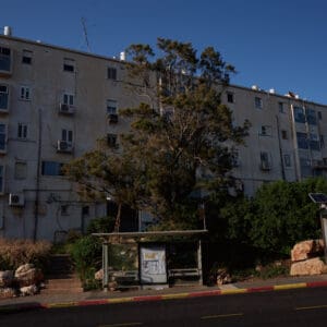 פינוי בינוי, התחדשות עירונית, תמ״א 38, הריסה ובנייה מחדש, חיזוק מבנים, שדרוג תשתיות, בנייה רוויה, צפיפות עירונית, תכנון עירוני, מדיניות דיור, התחדשות שכונות ותיקות, פיתוח עירוני, שינוי ייעוד קרקע, הגדלת זכויות בנייה, שימור מול פיתוח, חידוש חזיתות, שיפור איכות חיים, השקעות נדל״ן, עליית ערך נכסים, פרויקטים למגורים, שיתוף דיירים, פיצוי דיירים, יזמות נדל״ן, רגולציה תכנונית, היתרי בנייה, ועדות תכנון ובנייה, שיקום מבנים ישנים, התחדשות תשתיות עירוניות, התחדשות מרכזי ערים, בנייה לגובה, בניין מגורים ישן, שיכון, בלוק דירות, חזית בניין, ארכיטקטורה ישראלית, שכונת מגורים, עיר ישראלית, רחוב עירוני, תחנת אוטובוס, תחבורה ציבורית, תחנת המתנה, עץ אורן, צמחייה עירונית, מדרכה, אבני סלע, כביש רטוב, שמש חזקה, שמיים כחולים, אור וצל, חזית מוזנחת, תריסים סגורים, מזגנים חיצוניים, אנטנות על הגג, צילום רחוב, סביבה עירונית, דיור ציבורי, מבנה בטון, שכונה ותיקה, נוף עירוני