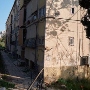 פינוי בינוי, התחדשות עירונית, תמ״א 38, הריסה ובנייה מחדש, חיזוק מבנים, שדרוג תשתיות, בנייה רוויה, צפיפות עירונית, תכנון עירוני, מדיניות דיור, התחדשות שכונות ותיקות, פיתוח עירוני, שינוי ייעוד קרקע, הגדלת זכויות בנייה, שימור מול פיתוח, חידוש חזיתות, שיפור איכות חיים, השקעות נדל״ן, עליית ערך נכסים, פרויקטים למגורים, שיתוף דיירים, פיצוי דיירים, יזמות נדל״ן, רגולציה תכנונית, היתרי בנייה, ועדות תכנון ובנייה, שיקום מבנים ישנים, התחדשות תשתיות עירוניות, התחדשות מרכזי ערים, בנייה לגובה, בניין מגורים ישן, שיכון, בלוק דירות, חזית בניין, ארכיטקטורה ישראלית, שכונת מגורים, עיר ישראלית, רחוב עירוני, תחנת אוטובוס, תחבורה ציבורית, תחנת המתנה, עץ אורן, צמחייה עירונית, מדרכה, אבני סלע, כביש רטוב, שמש חזקה, שמיים כחולים, אור וצל, חזית מוזנחת, תריסים סגורים, מזגנים חיצוניים, אנטנות על הגג, צילום רחוב, סביבה עירונית, דיור ציבורי, מבנה בטון, שכונה ותיקה, נוף עירוני