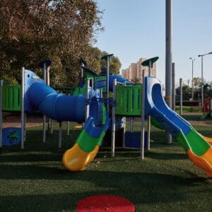 גן משחקים, מתקני משחק, מגלשה כחולה, מגלשה צהובה, מנהרת זחילה, פארק עירוני, דשא סינתטי, בטיחות ילדים, ציוד שעשועים, חצר ציבורית, אזור פנאי, משחקי ילדים, מתקן טיפוס, עמודי תאורה, עצים ירוקים, ספסלי ישיבה, בנייני מגורים ברקע, יום שמשי, צללים ארוכים, סביבה עירונית, פארק שכונתי, חוץ, צבעוני, ריק מאנשים, תשתית עירונית
