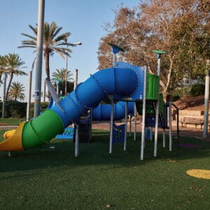 גן משחקים, מתקני משחק, מגלשה כחולה, מגלשה צהובה, מנהרת זחילה, פארק עירוני, דשא סינתטי, בטיחות ילדים, ציוד שעשועים, חצר ציבורית, אזור פנאי, משחקי ילדים, מתקן טיפוס, עמודי תאורה, עצים ירוקים, ספסלי ישיבה, בנייני מגורים ברקע, יום שמשי, צללים ארוכים, סביבה עירונית, פארק שכונתי, חוץ, צבעוני, ריק מאנשים, תשתית עירונית