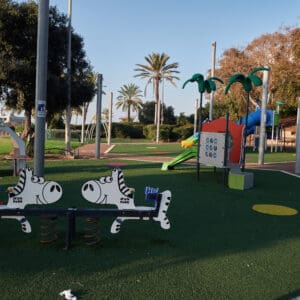 גן משחקים, מתקני משחק, מגלשה כחולה, מגלשה צהובה, מנהרת זחילה, פארק עירוני, דשא סינתטי, בטיחות ילדים, ציוד שעשועים, חצר ציבורית, אזור פנאי, משחקי ילדים, מתקן טיפוס, עמודי תאורה, עצים ירוקים, ספסלי ישיבה, בנייני מגורים ברקע, יום שמשי, צללים ארוכים, סביבה עירונית, פארק שכונתי, חוץ, צבעוני, ריק מאנשים, תשתית עירונית, גן אלכס
