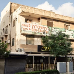 רחוב הרצל, הדר הכרמל, מסחר, חנויות, תריסי גלילה, מדרכה, ריצוף, כביש, שלטי רחוב, תמרורים, מגדל חיפה, בנייני מגורים, צמחייה עירונית, עצי סיגלון, פריחה סגולה, בובת תצוגה, פח אשפה, גרפיטי, שלטי השכרה, נוף עירוני, תשתיות, מבנה מוזנח, הזנחה, ישן, התרוקנות, עזיבה, ג׳נטריפיקציה, מבנה נטוש, נטישה, נטושים