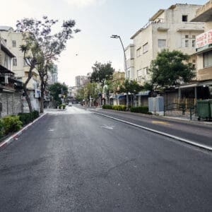 רחוב הרצל, הדר הכרמל, מסחר, חנויות, תריסי גלילה, מדרכה, ריצוף, כביש, שלטי רחוב, תמרורים, מגדל חיפה, בנייני מגורים, צמחייה עירונית, עצי סיגלון, פריחה סגולה, בובת תצוגה, פח אשפה, גרפיטי, שלטי השכרה, נוף עירוני, תשתיות