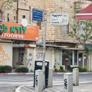 דודא לפיצה, פיצריה, משלוח, שילוט בעברית, רחוב הרצל, רחוב יחיאל, הדר הכרמל, מסחר, חנויות, תריסי גלילה, מדרכה, ריצוף, כביש, שלטי רחוב, תמרורים, מגדל חיפה, בנייני מגורים, צמחייה עירונית, עצי סיגלון, פריחה סגולה, בובת תצוגה, פח אשפה, גרפיטי, שלטי השכרה, נוף עירוני, תשתיות