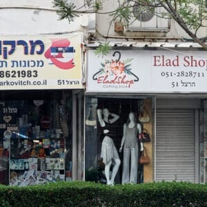 מסחר, חנויות, רחוב הרצל, הדר הכרמל, חזית עסק, תריסי גלילה, שלטים, חנות בגדים, חנות תכשיטים, מכונות תפירה, מדרכה, ריצוף אבן, בובות תצוגה, חלון ראווה, גרפיטי, אמנות רחוב, סמטה, פח אשפה ירוק, צמחייה עירונית, שיחים, עמודי תאורה, תמרורים, מרקוביץ׳ מכונות תפירה, אלעד שופ, ELAD SHOP