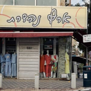 מסחר, חנויות, רחוב הרצל, הדר הכרמל, חזית עסק, תריסי גלילה, שלטים, חנות בגדים, חנות תכשיטים, מכונות תפירה, מדרכה, ריצוף אבן, בובות תצוגה, חלון ראווה, גרפיטי, אמנות רחוב, סמטה, פח אשפה ירוק, צמחייה עירונית, שיחים, עמודי תאורה, תמרורים, ארץ נהדרת, שילוט בעברית
