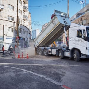משאית, משאית רכינה, פריקה, עבודות בכביש, פועלים, אתר בנייה, כביש, אספלט, סופר פארם, בניינים, אדריכלות, חול, אבק, גדר בטיחות, קונוסים, תשתיות, רחוב עירוני, פיתוח, בנייה, תחבורה, ציוד מכני הנדסי, אפוד זוהר